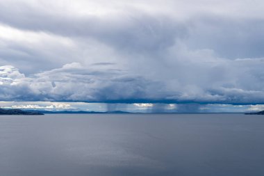 Taquile Adası 'ndan Titicaca Gölü' nün geniş manzarası. Durgun sular, dalgalanan beyaz ve mavi bulutlarla dolu dramatik bir gökyüzünü yansıtıyor.