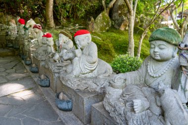 Kırmızı örgü şapkalar ve önlükler giyen bir grup yosun kaplı Jizo Bosatsu heykeli Daisho-in Tapınağı, Miyajima 'da gölgeli bir orman yolu çiziyor.