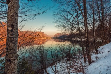 Beautiful sunset scenic image of Lake Bohinj and old Slovenian village Stara Fuzina in background seen through branches of deciduous trees by the water