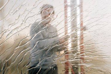 Man washing car with water gun in carwash self-service. Soap sud, wax and water drops covering vehicle window glass. Seen from the inside of the automobile.