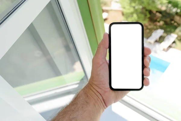 スマートフォンのモックアップ画面 夏に屋外スイミングプールのビューとアパートの窓の横に空白のタッチスクリーンを持つ携帯電話を保持男 選択的フォーカス — ストック写真