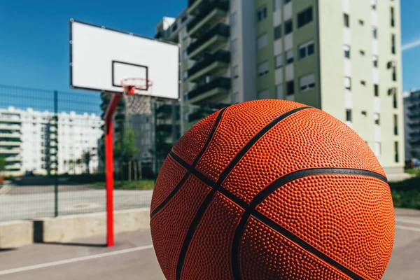 Basketball ball on outdoor court with concrete surface and backboard with hoops in background in residential district, selective focus