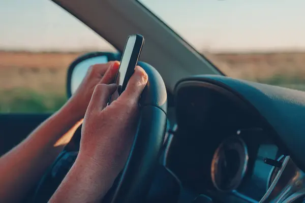 車を運転しながらテキストメッセージングに携帯電話を使用する女性 スマートフォンと車両のステアリングホイールを保持する女性の手のクローズアップ 選択的な焦点 — ストック写真