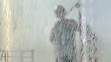 Man washing car with water gun in carwash self-service. Soap sud, wax and water drops covering vehicle window glass. Seen from the inside of the automobile.