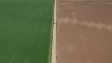 Güneşli bir bahar gününde kırsal alanda araba sürmek, düz yolda düz arazide siyah arabanın havadan çekilmiş görüntüsü insansız hava aracı pov 'undan