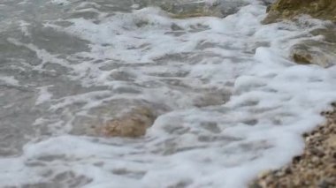 Yavaş çekim dalgaları ve deniz köpüğü yaz sabahı çakıl taşlı plajı eziyor, seçici bir odak noktası var.