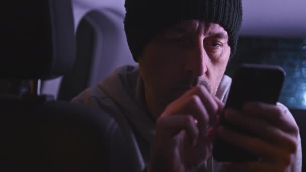 夜の車の後部座席で携帯電話を使用している男性 黒い帽子と灰色のフード付きの大人の男性の低いキー映像 選択的な焦点 — ストック動画