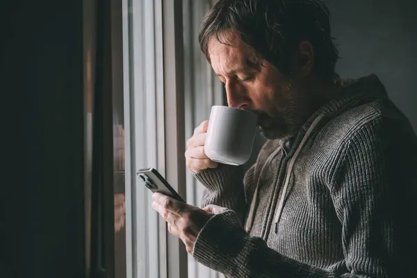 早上喝咖啡休息的时候 一个头脑发热的成年人从白色的杯子里喝着咖啡 一边在社交媒体上浏览着他的智能手机 一边站在公寓的窗边 有选择地集中精神 — 图库照片