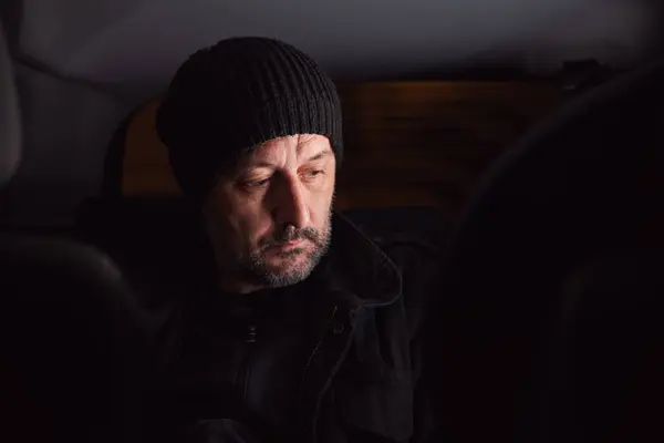 Serious worried man thinking at back seat of the car at night. Adult male wearing black cap and jacket and thinking. Selective focus.