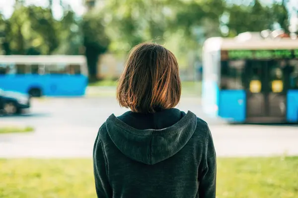 stock image Rear view of adult brunette female standing on the street and looking at traffic and buses passing by, selective focus