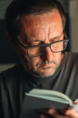 Kitap kurdu konsepti. Evde kitap okumak, gecenin geç saatlerinde yalnız başına yetişkin bir erkeğin gösterişsiz portresi, seçici bir odak noktası..