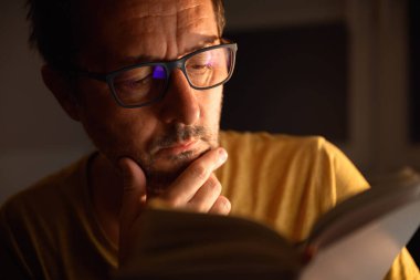Kitap kurdu karanlık odada edebiyat kitabı okuyor, yüzü masa lambası ışığıyla aydınlanıyor. Seçici odak.