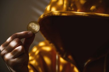Bilgisayar korsanı ve kripto para dolandırıcısı altın sarısı kapüşonlu ceketli elinde Bitcoin, seçici odaklı