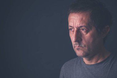 Karanlık odada gri tişört giyen, ciddi bir orta yaşlı erkek portresi.