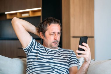 Evde tek başına, rahat kıyafetlerle kanepede oturan ve elinde akıllı telefon tutan sıkıcı bir adam. Seçici odak.