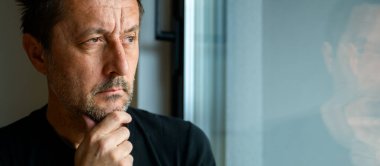 Ciddi bir beyaz yetişkin erkek pencereden bakıyor ve düşünüyor, seçici odaklanıyor.