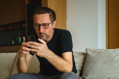 Evde tek başına, rahat kıyafetlerle kanepede oturan ve elinde akıllı telefon tutan sıkıcı bir adam. Seçici odak.