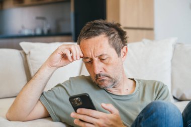 Evde tek başına, rahat kıyafetlerle kanepede oturan ve elinde akıllı telefon tutan sıkıcı bir adam. Seçici odak.