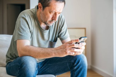 Evde tek başına, rahat kıyafetlerle kanepede oturan ve elinde akıllı telefon tutan sıkıcı bir adam. Seçici odak.