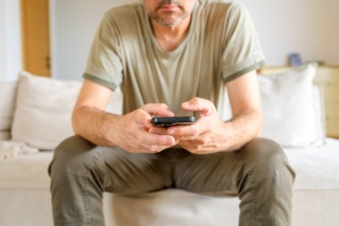 Kanepede sıradan kıyafetlerle oturan bir adam akıllı telefon tutuyor. Seçici odak.
