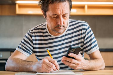 Adam kağıt, kalem ve akıllı telefon hesap makinesi kullanarak ev bütçesi hesaplaması yapıyor.