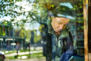 Genel kütüphane rafında kitaplara göz atan sıradan bir kadının portresi, geniş pencereden görülüyor, seçici odak noktası