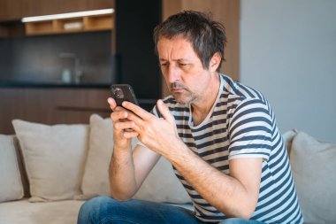 Evde tek başına, rahat kıyafetlerle kanepede oturan ve elinde akıllı telefon tutan sıkıcı bir adam. Seçici odak.