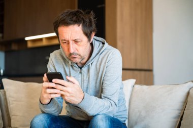 Evde tek başına, rahat kıyafetlerle kanepede oturan ve elinde akıllı telefon tutan sıkıcı bir adam. Seçici odak.