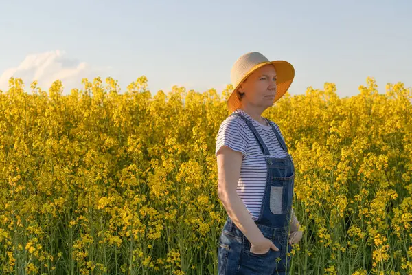 Güneşli bir bahar öğleden sonrasında çiçek açan kanola tarlasında hasır şapkalı güzel bir kadın çiftçinin portresi, seçici bir odak noktası.