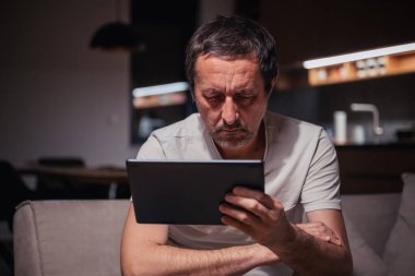 Gecenin geç saatlerinde evde dijital tablet bilgisayardan sosyal medya içeriği okuyan yorgun adam, seçici odaklanma
