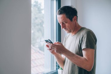 Evde pencerenin yanında cep telefonu kullanan sıradan bir yetişkin adam, seçici odaklı.