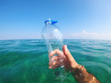 Okyanus kirliliğini ve çevresel farkındalığı ön plana çıkaran, denizde boş bir plastik şişe tutan el. Mavi gökyüzünün altında turkuaz bir suyun içinde vuruldu. Seçici odak.