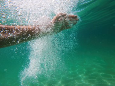 Suyun altındaki bir el, berrak bir turkuaz denizde su sıçratır ve kabarcıklar oluşturur. Güneş ışığı da suyun içinden süzülür. Seçici odak.