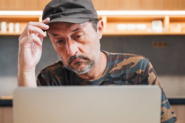Kamuflaj gömleği ve asker şapkası giyen adam evde dizüstü bilgisayar kullanıyor, seçici odaklı.