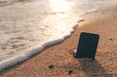 Cep telefonu gün batımında köpüklü deniz dalgası yakınlarında kumlara sıkışmış. Risk, kaza ya da plajda kayıp kavramı. Seçici odak.