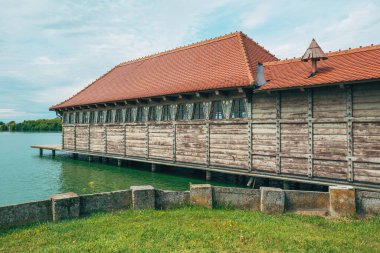 Sırbistan 'ın Paliç Gölü' nde parlak bir yaz gökyüzü altında sakin yeşil su üzerinde uzanan süslü ayrıntılara sahip geleneksel ahşap kayıkhane. Seçici odak