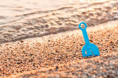 Plajda kumlara saplanmış mavi plastik bir oyuncak küreğe yakın plan, arka planda hafif dalgalar ve altın güneş ışığı var. Yaz mevsimi, çocukluk ve plaj zamanı kavramı. Seçici odak.