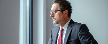 Concerned executive in formal attire gazing through office window, reflecting on challenges. Selective focus.