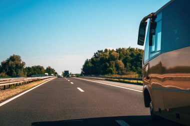 Golden bus on the highway like its racing toward vacation dreams and holiday escape. Side view of a bus driving on road on sunny day with traffic and trees.