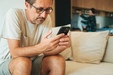 Middle-aged man sitting on a sofa using smartphone while woman works in the kitchen in the background. Selective focus.