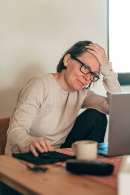 Disappointed female freelancer with hand on head looking at laptop screen in home office workspace. Selective focus.
