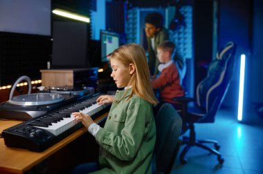 Okul çocukları ses kayıt stüdyosunda müzik yapıyor, sentezleyici oynayan kıza odaklanıyor.
