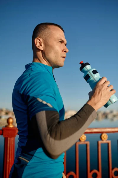 Portrait of healthy athletic middle aged man with fit body holding bottle of refreshing water, resting after workout or running. Male runner drink after outdoor training