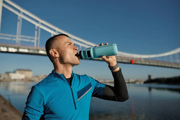 Portrait of healthy athletic middle aged man with fit body drinking water, resting after workout or running. Male runner drink after outdoor training against bridge