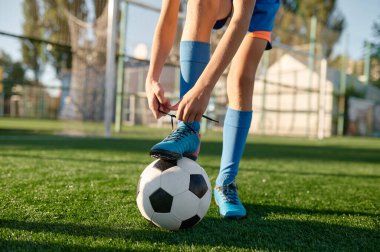 Ayakkabı bağcıkları bağlayan küçük futbolcu futbol topuna bacak koyuyor. Spor okulunda antrenman için hazırlık