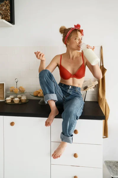 台所のテーブルに座りながら瓶から牛乳を飲むランジェリー姿の若い女性 健康的なライフスタイルと幸せな朝 — ストック写真