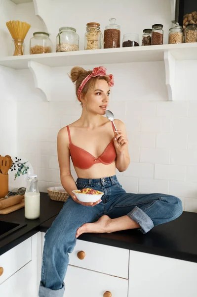 ランジェリーブラジャーとジーンズのレトロなスタイルの若い女性が自宅のキッチンテーブルの上に座ってミルクでフレークを食べています 朝のトレーニング後の食事と健康的な朝食 — ストック写真