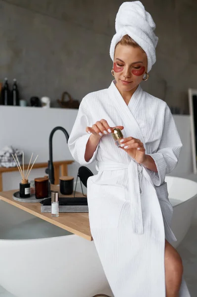 Young woman in towel applying cosmetic beauty patch under eyes to remove wrinkles after taking bath or shower. Skin care, beauty treatment, cosmetology, dermatology concept