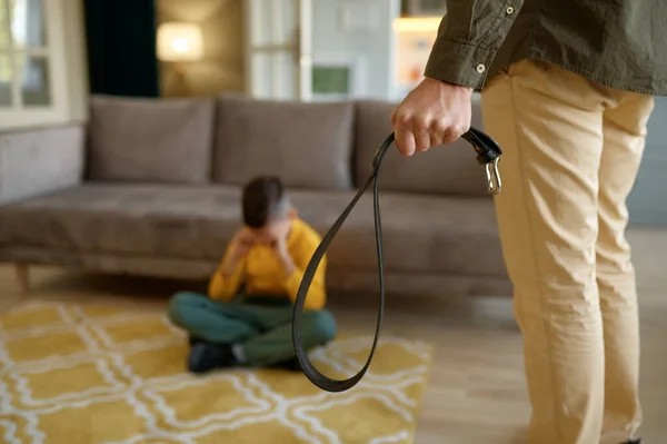 Closeup of father holding leather belt to punish crying afraid son sitting on floor. Parental abuse, bad relationship, violence in family concept