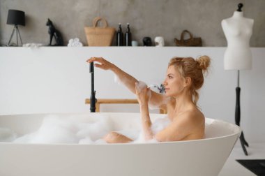 Genç bir kadın ev banyosunda köpük köpükleriyle banyo yapıyor. Milenyum dişisi küvette dinleniyor tatilin ve hafta sonunun keyfini çıkarmanın hayalini kuruyor.